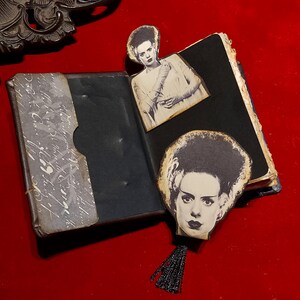 May include: An open, vintage-style book with black pages and a decorative cover. The book features two black and white images of the Bride of Frankenstein, one as a bookmark with a black tassel. The background is a deep red.