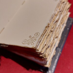 May include: Open journal with aged, textured pages. The pages are off-white with deckled edges. Gold filigree corner designs adorn the pages. The journal's spine is dark blue-grey. The background is a solid red.