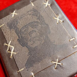 May include: A brown leather wallet with a stitched-on square featuring a black and white image of a person. The square is stitched to the wallet with cream-colored thread in an X pattern. The wallet is rectangular.