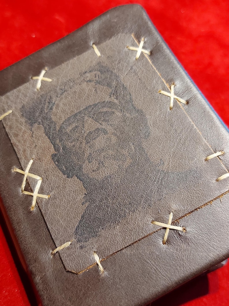 May include: A brown leather wallet with a stitched-on square featuring a black and white image of a person. The square is stitched to the wallet with cream-colored thread in an X pattern. The wallet is rectangular.