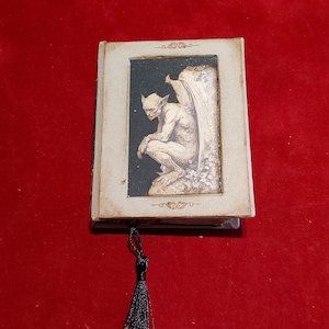 Puede incluir: Un libro rectangular con una cubierta de color crema que presenta una ilustración central incrustada de una figura de gárgola alada en una pose sentada y contemplativa. El libro incluye un marcapáginas de borla de tela negra.
