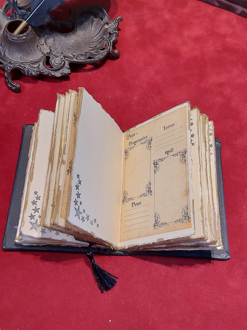 Puede incluir: Un libro abierto de estilo antiguo con p&aacute;ginas envejecidas de color crema. Las p&aacute;ginas est&aacute;n decoradas con dise&ntilde;os de estrellas y flores. El libro tiene una cubierta negra y una borla negra. El texto en las p&aacute;ginas incluye las palabras "Date", "Preparation", "Intent", "Spell" y "Notes".