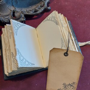 Puede incluir: Un peque&ntilde;o diario de cuero negro con bordes decorativos y una etiqueta en blanco adjunta. El diario tiene una est&eacute;tica vintage y est&aacute; abierto en una p&aacute;gina en blanco.