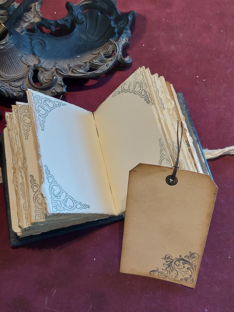 Puede incluir: Un peque&ntilde;o diario de cuero negro con bordes decorativos y una etiqueta en blanco adjunta. El diario tiene una est&eacute;tica vintage y est&aacute; abierto en una p&aacute;gina en blanco.