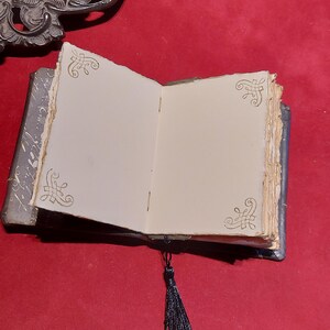 May include: An open, antique-style journal with blank, off-white pages. The journal has a dark blue cover and a black tassel. Decorative gold filigree accents the corners of the pages. The journal is on a red surface.
