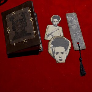 May include: A dark brown book with a Frankenstein image and stitched accents, alongside two Bride of Frankenstein cutouts and a bookmark with a black tassel. The items are arranged on a red surface.