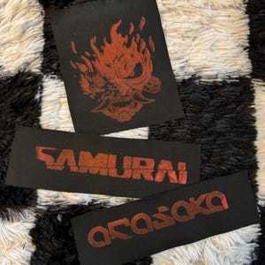May include: Three black fabric patches with red designs. One patch features a flaming skull, while the others display the words "SAMURAI" and "ARASAKA" in a stylized font. The patches are likely for clothing or accessories.