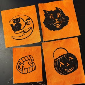 May include: Four orange fabric squares with black Halloween-themed designs. The designs include a cat on a crescent moon, a grinning cat face, vampire teeth, and a jack-o'-lantern bucket. The squares are arranged on a dark surface.