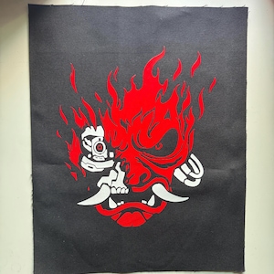 May include: A rectangular black fabric patch featuring a graphic design of a stylized robotic samurai mask. The design is rendered in vibrant red and white, depicting a mechanical face with sharp fangs, flames, and intricate cybernetic details.