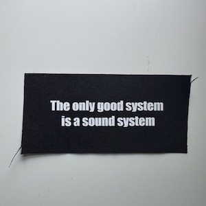 May include: A rectangular black fabric patch with white text printed in the center that reads, The only good system is a sound system. The patch has a woven texture and visible loose threads at the corners, indicating it is a sew-on garment accessory.