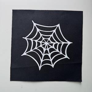 May include: A square black fabric patch featuring a centered white graphic print of a spider web. The design consists of concentric lines connected by radiating spokes, creating a symmetrical web pattern against the solid black textile background.