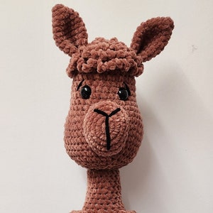 Op de afbeelding: Een bruine gehaakte lama met een kleinere groene gehaakte lama die op zijn schoot zit. Beide lama's hebben zwarte ogen en neuzen.