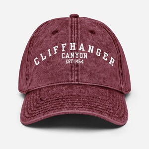 Puede incluir: Gorra de béisbol granate con el texto "CLIFFHANGER CANYON EST 1454" en letras blancas. La gorra tiene una visera curva y un cierre de correa de tela. El sombrero está hecho de un material suave y desgastado.