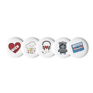 May include: Set of five white round buttons with colorful illustrations. The buttons feature a broken heart with a bandage, a sad cloud with raindrops and lightning, headphones with a broken heart, a blue bear wearing a hoodie, and a blue cassette tape with the text "Still Not Okay!"