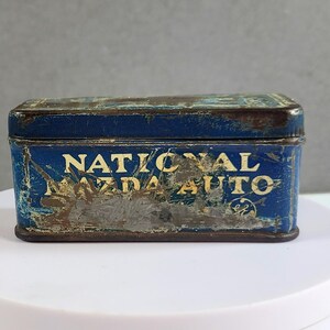 May include: A rectangular, vintage blue tin with the words "NATIONAL MAZDA AUTO" in gold lettering. The tin shows signs of wear and tear, with rust and paint loss, suggesting age. The overall aesthetic is antique.