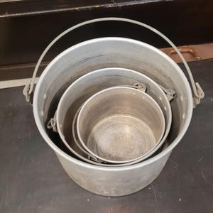 May include: Three nested aluminum buckets with attached handles. The buckets are different sizes, with the largest bucket on the outside and the smallest bucket nested inside. The buckets are silver in color and appear to be vintage.