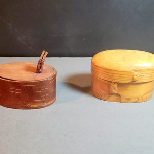 May include: Two wooden boxes. One is cylindrical with a lid and a twig handle, the other is oval with a hinged lid. Both boxes are made of natural wood and have a rustic appearance. The boxes are likely used for storage or decoration.