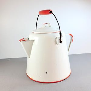 May include: White enamelware tea kettle with a red handle and trim. The kettle has a spout, a lid, and a black wire handle. The red trim is visible around the base, spout, and lid. The kettle is a classic design.