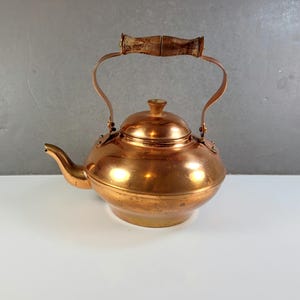 May include: A copper tea kettle with a wooden handle and a curved spout. The kettle has a round body and a lid with a small knob. The handle is made of wood and is attached to the kettle with copper brackets.