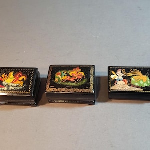 May include: Three black lacquered boxes with intricate, colorful scenes. The boxes feature gold trim and detailed illustrations of figures and animals. The boxes are rectangular and appear to be small trinket boxes.