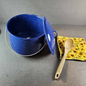 May include: A deep blue enamel pot with a speckled finish, a metal bail handle, and a matching lid. Beside the pot lies a light-colored wooden spoon resting on a square quilted potholder featuring a vibrant yellow sunflower pattern.