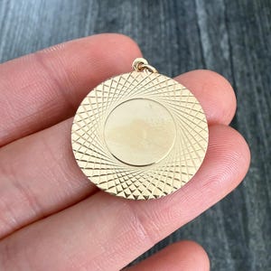 May include: A gold-colored circular pendant with a textured spiral design around a smooth center. The pendant is held in a hand, showcasing its size and detail. The pendant has a small loop at the top for a chain.