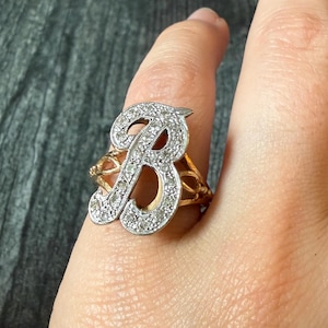 Könnte beinhalten: Ein Goldring mit einem großen, markanten Buchstaben B als Herzstück. Der Buchstabe B ist mit zahlreichen kleinen, klaren, funkelnden Steinen besetzt, die in silberfarbenes Metall gefasst sind, während die Ringschiene aus poliertem Gelbgold besteht.