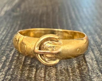 May include: A gold-colored ring with a buckle design. The ring has a rounded band and a detailed buckle clasp. The metal has a slightly aged appearance, suggesting it may be vintage or antique. The ring is displayed on a textured surface.