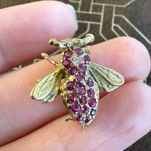 Puede incluir: Un broche de abeja de oro con detalles de rubíes y diamantes. La abeja está detallada con alas y un cuerpo cubierto de rubíes rojos.