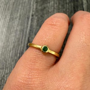 May include: A gold ring with a small, round, emerald-green gemstone set in the center. The ring is simple in design, with a thin band and a bezel setting for the gemstone. The ring is worn on a finger.
