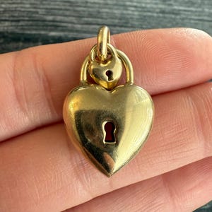 Könnte beinhalten: Ein goldfarbener herzförmiger Anhänger mit Schlüsselloch-Design. Der Anhänger hat ein kleineres herzförmiges Element oben, ebenfalls mit einem Schlüsselloch. Der Anhänger wird in einer Hand gehalten.