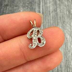 May include: A small, ornate pendant in the shape of the letter "B". The pendant is silver-toned with small, clear stones and a gold-colored bail. The jewelry is held between fingers against a blurred background.