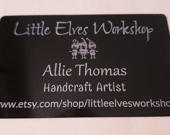 May include: Black business card with white text that reads "Little Elves Workshop" with a small image of three elves holding hands. The text "Allie Thomas Handcraft Artist" is below the image. The website address "www.etsy.com/shop/littleelvesworkshop" is at the bottom of the card.