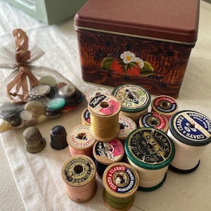 May include: A collection of sewing supplies including spools of thread, thimbles, and a decorative tin. The spools are in various colors, and the tin features a strawberry and flower design. A small bag of buttons is also present.
