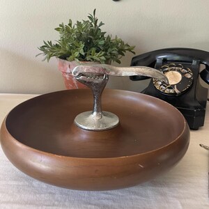 May include: A round, dark brown wooden bowl featuring a central, integrated silver-toned metal nutcracker mechanism. The bowl has a smooth, curved interior and exterior surface, with the metallic nutcracker rising from the center of the base.