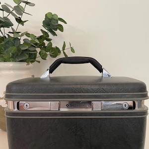 May include: A vintage Samsonite black travel case with a black handle and silver-toned accents. The case has two silver latches and the Samsonite logo. The case is sitting on a white surface.