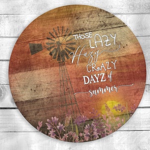 May include: A round wooden sign with a rustic woodgrain background. The sign features a silhouette of a windmill with a sunset in the background. The text on the sign reads "Those Lazy Hazy Crazy Dayz of Summer".