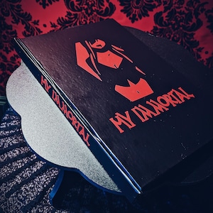 Puede incluir: Un libro negro con el título "MY IMMORTAL" en letras rojas. La portada presenta un gráfico rojo de una persona. El libro descansa sobre un soporte plateado y azul, con un fondo rojo estampado.