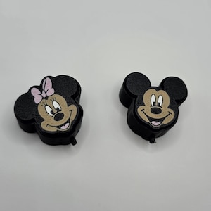 May include: Two black plastic badge reels shaped like the heads of iconic cartoon characters. One features a character with a light pink bow, while the other features a character with a tan face. Both have textured black ears and a small black hook.
