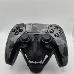 May include: A camouflage-patterned PlayStation 5 controller rests on a black, stylized Oni mask stand. The controller features a gray and black camo design on the buttons and handles, with the PlayStation logo visible. The mask stand adds a unique aesthetic.
