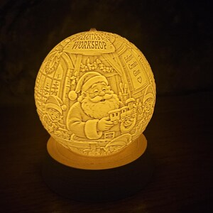 May include: A spherical lamp with a warm, glowing light featuring a detailed relief carving of Santa Claus holding a toy train inside a room labeled SANTA'S WORKSHOP. The textured, off-white globe sits securely on a circular wooden base.