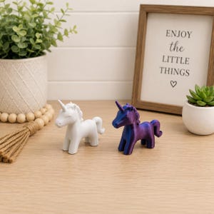 May include: Two small unicorn figurines standing side by side. One unicorn is solid matte white, while the other features a metallic, iridescent finish that shifts between deep purple and blue tones. Both figures have a horn and a sculpted mane.