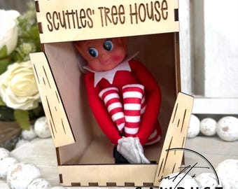 May include: A wooden box with the text "Scuttles' Tree House" houses a red and white elf doll. The doll is wearing a red and white striped outfit and is sitting inside the box. The box has a natural wood finish and a jute string handle.