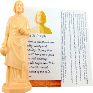 May include: A light peach-colored Saint Joseph statue, holding a jug, stands next to a card with instructions. The card features an image of Saint Joseph and text about selling a house. The statue is on a small base.