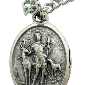 May include: Silver oval pendant necklace with a depiction of Saint Hubert, a patron saint of hunters, with a stag and a dog. The pendant is on a silver chain.