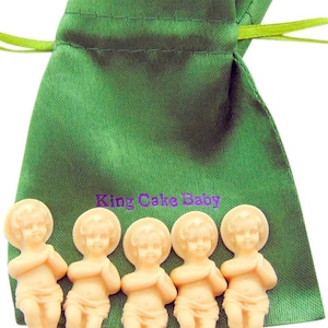 May include: A green satin drawstring bag with the text "King Cake Baby" in purple. Five small, light-colored plastic baby figurines are in front of the bag. The babies have their hands clasped in prayer.
