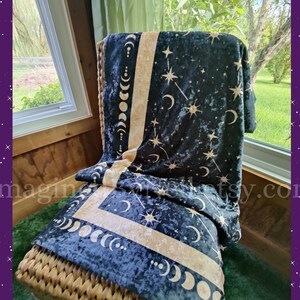 May include: A dark blue blanket with a celestial design of moons, stars, and constellations in a gold color. The blanket is draped over a wicker chair, creating a cozy and inviting scene. The blanket's design is repeated on the border.
