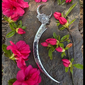 Puede incluir: Un peine de plata en forma de media luna con un diseño de fases lunares y un patrón de estrellas. El peine está decorado con una flor y una piedra transparente en el centro. El peine está rodeado de flores rosas.