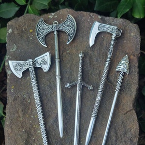 May include: A collection of silver-colored miniature weapons, including axes, a sword, and an arrow. The axes feature intricate Celtic knot designs. The items are displayed on a rough, brown stone surface.