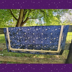 May include: A dark blue velvet tapestry with a celestial design of stars, moons, and shooting stars. The tapestry has a gold border with moon phases. The fabric is draped over a wooden fence, with a green background.